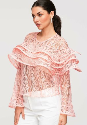 Femme aux cheveux foncés attachés en queue de cheval, portant des boucles d'oreilles pendantes et un chemisier en dentelle rose transparent avec des volants superposés, associé à un pantalon blanc taille haute.