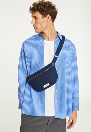 Chemise à boutons à rayures bleues et blanches portée par-dessus un t-shirt blanc, avec une banane en tissu bleu foncé dotée d'une fermeture éclair et d'un logo.