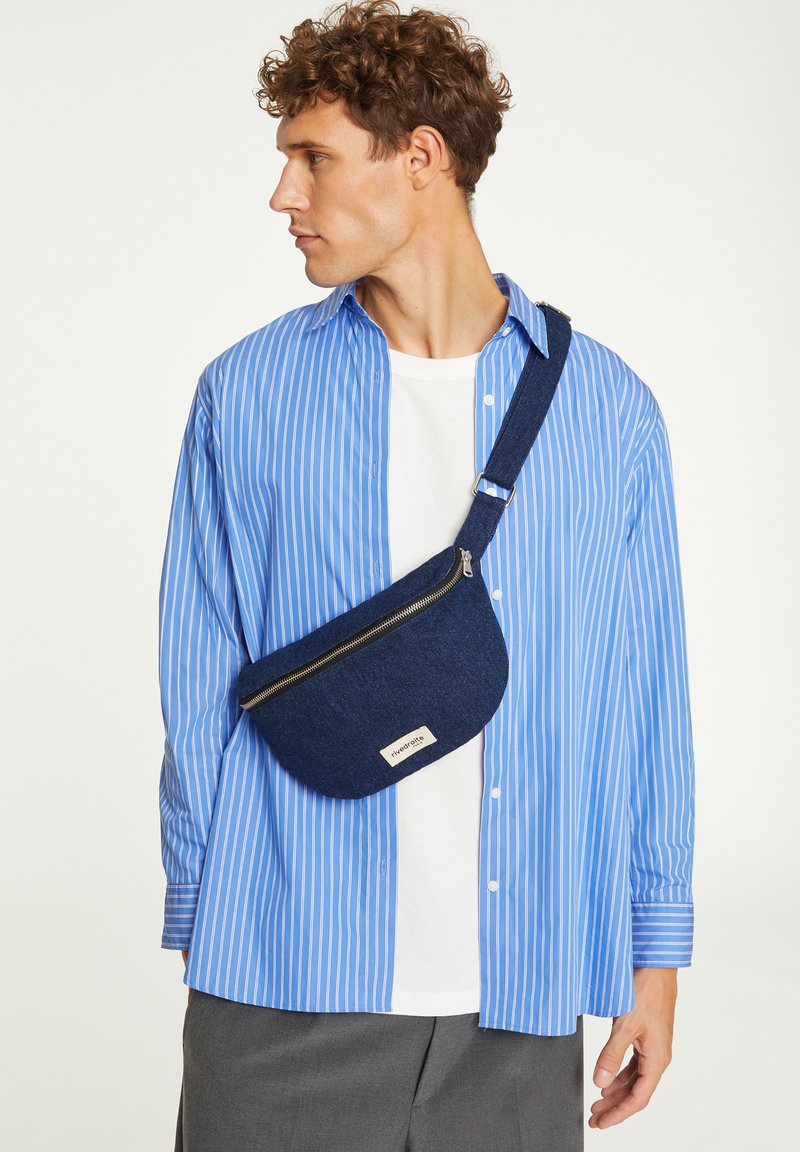 Chemise à boutons à rayures bleues et blanches portée par-dessus un t-shirt blanc, avec une banane en tissu bleu foncé dotée d'une fermeture éclair et d'un logo.