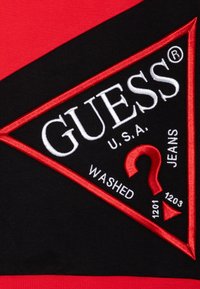 Zwarte driehoekige Guess-logo patch met rode rand en tekst "GUESS U.S.A. WAShed JEANS" en een rode vraagteken op een rood en zwart stoffen achtergrond.