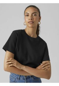 T-shirt in cotone nero con maniche corte e scollo rotondo, indossato con le braccia incrociate. Jeans visibili, abbigliamento casual con texture.