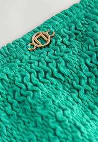 Teal värviline kootud kangas, millel on tekstuuriga laine muster, millel on kuldtooniline ümmargune klamber. Suurendatud vaade, mis toob esile kanga detailid.