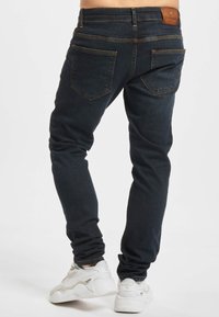 Tmavě modré džíny s úzkým střihem, vybavené dvěma zadními kapsami, žlutým prošitím a hnědou koženou nášivkou na pasu. Kombinovány s bílými teniskami.