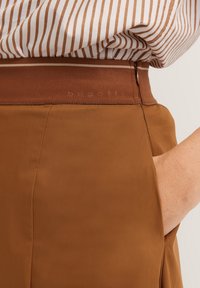 Braune Hose mit glatter Textur, ausgestattet mit einem breiten Bund und Seitentaschen. Gestreiftes Hemd ist teilweise sichtbar.