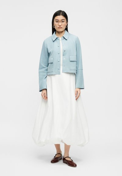 Moteris su ilgais tamsiais plaukais ir akiniais, dėvinti šviesiai mėlyną užsegamą švarką, baltą čiurnos ilgio suknelę ir rudus Mary Jane stiliaus batus.