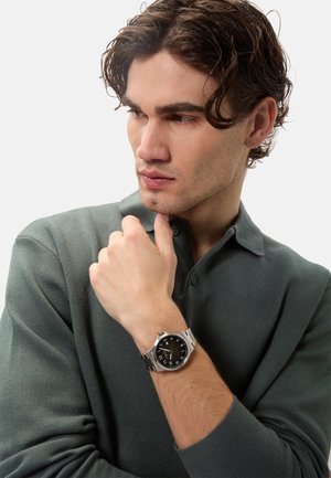 Orologio da uomo con quadrante nero, cinturino in acciaio inossidabile argentato e cassa rotonda. Indossato su un polso, presenta marcatori delle ore chiari e una texture liscia.