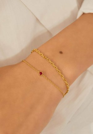 Pols met twee gouden armbanden: een dunne ketting met een kleine rode edelsteen en een dikkere schakelkettinkje.