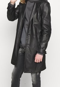 Abrigo de cuero negro con un diseño elegante, cierre de un solo botón y grandes bolsillos. Lleva puesto un sudadera gris con capucha; cuenta con puños enrollados.