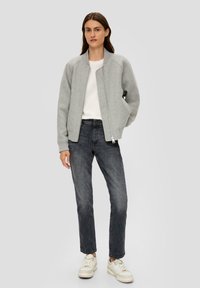 Veste bomber grise avec des poignets côtelés, fermeture éclair, portée sur un t-shirt blanc. Assortie à un jean noir skinny et des baskets claires. Design simple.