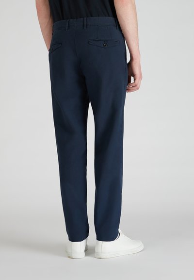 Pantalons bleu marine en tissu doux, avec un devant plat, deux poches arrière avec boutons, et une coupe droite, associés à des chaussures blanches.