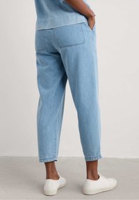 Light blue denim trousers with a relaxed fit, tapered legs, side pockets, and a clean hem. Worn with white slip-on shoes.