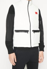 Weißer Puffer-Weste mit hohem Kragen, schwarzen Ärmeln und Reißverschlusstaschen. Mit einem auffälligen roten Logo-Patch auf der Brust.