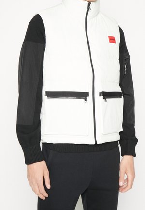 Witte gewatteerde vest met hoge kraag, zwarte mouwen en ritszakken. Bevat een opvallende rode logo-patch op de borst.