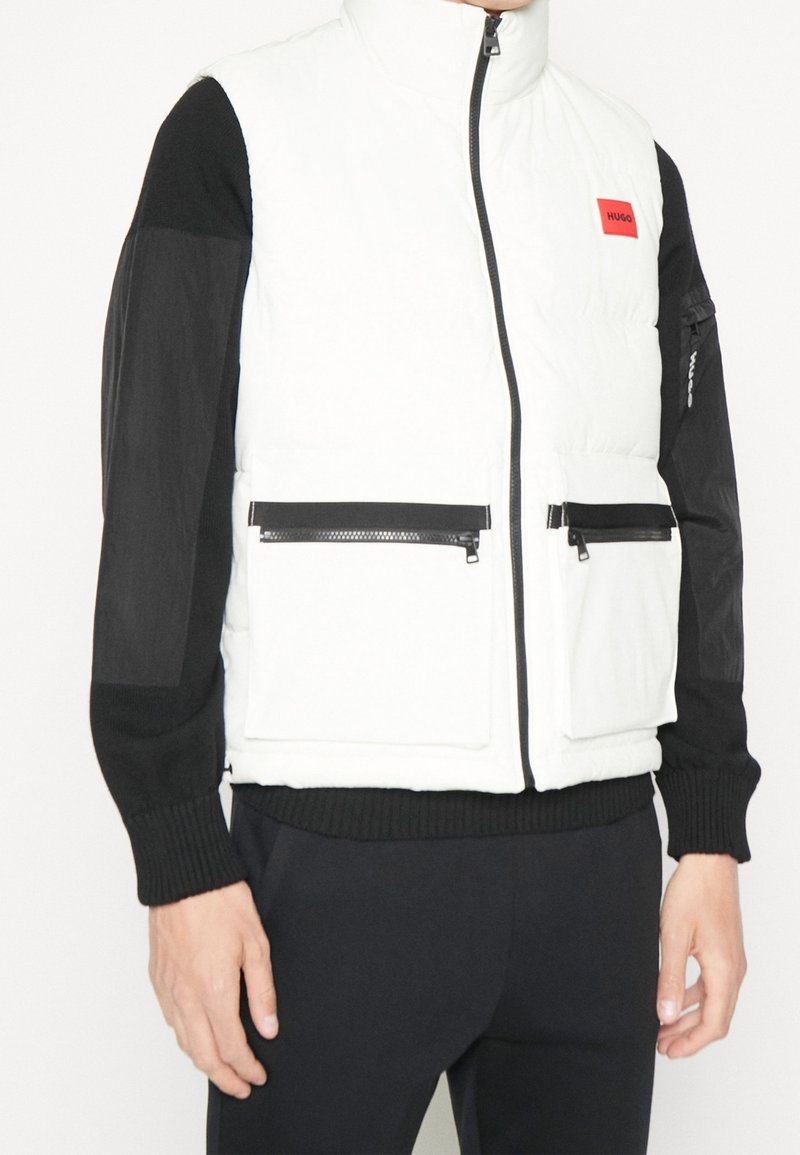 Weißer Puffer-Weste mit hohem Kragen, schwarzen Ärmeln und Reißverschlusstaschen. Mit einem auffälligen roten Logo-Patch auf der Brust.