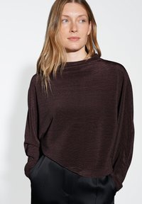 Frau mit langen blonden Haaren, die ein dunkles geripptes Langarmoberteil und schwarze Hosen trägt, mit den Händen in den Taschen, und zur Seite schaut.