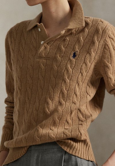 Személy, aki tevés színű, fonott mintás kötött pulóvert visel gallérral és kék hímzett logóval, begyűrve szürke nadrágba.