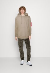 Svetlo rjava kapuca s prednjim žepom in oznako na roki, v kombinaciji z olivno zelenimi kordnimi hlačami in belimi supergami.