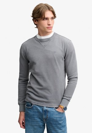 Ung mand med lyst brunt hår iført en grå langærmet skjorte over en hvid undertrøje og blå jeans, kigger til siden med den ene hånd i lommen.