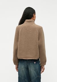 Chaqueta de forro polar marrón con cuello alto, mangas raglán y un corte relajado. Textura suave con un dobladillo elástico. Llevada sobre un vaquero.