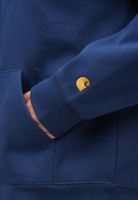 Tamsiai mėlynas džemperis su priekine kišene, ribinėmis rankogaliais ir geltonu siuvinėtu bangos logotipu kairėje rankovėje. Minkštas, faktūruotas audinys.