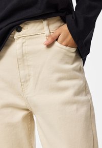 Mano con smalto neutro appoggiata nella tasca anteriore di pantaloni beige chiaro, indossati con una maglia nera a maniche lunghe.