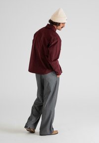 Hombre con un gorro de punto color crema, chaqueta burdeos, pantalones de pierna amplia a rayas grises y zapatos color beige, de pie con las manos en los bolsillos, mirando ligeramente hacia abajo y a la derecha.