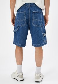 Pantalones cortos de cargo de mezclilla en azul medio con un ajuste relajado, que presentan dos grandes bolsillos traseros y un bolsillo lateral de cargo con un parche de logo.