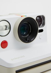 Appareil photo Polaroid blanc avec un objectif rond noir, un bouton rouge et un design à rayures arc-en-ciel. La surface texturée et l'étiquetage de mise au point automatique sont visibles.