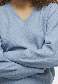 Pull en tricot bleu clair avec un col en V et un motif chevron texturé. Manches longues et poignets côtelés. Matière douce et chaude.