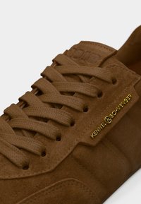 Καφέ δερμάτινο αθλητικό παπούτσι με υφή, χρυσό λογότυπο και επίπεδα κορδόνια. Διαθέτει χαμηλή σχεδίαση και ενισχυμένες τρύπες για τα κορδόνια.