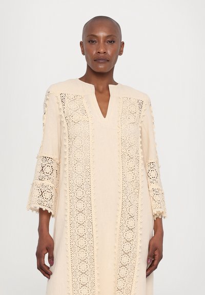 Beige tunic with a V-neck, featuring intricate lace detailing and 3/4 sleeves. Soft texture with decorative pom-pom accents along the edges.