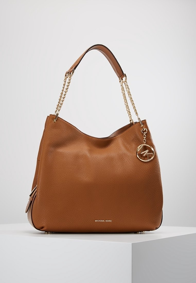 Sac à main en cuir brun à texture granuleuse, avec un design décontracté, des anses en chaîne dorée et un charm en forme de logo circulaire doré attaché à l'avant.