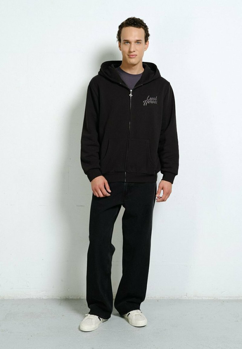 Junger Mann steht vor einer weißen Wand und trägt einen schwarzen Zip-Hoodie mit dem Text "Local Heroes", schwarze Hosen und weiße Sneakers.