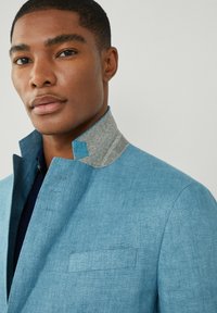 Blazer azzurro chiaro con tessuto testurizzato, caratterizzato da un colletto grigio e un distintivo accento blu sul bordo del colletto, e una singola tasca sul petto.