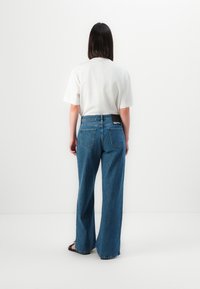 Φαρδιά μπλε τζιν παντελόνια με ακατέργαστο στρίφωμα, διαθέτουν πίσω τσέπες και λογότυπο στη μέση; συνδυασμένα με ένα απλό λευκό μπλουζάκι.