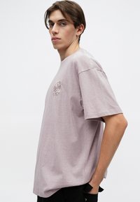 Απαλό μωβ βαμβακερό T-shirt με άνετη εφαρμογή, στρογγυλή λαιμόκοψη και κοντά μανίκια. Διαθέτει κεντητό λογότυπο στο στήθος. Λείο υλικό.