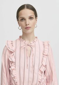 Rožinė bluzonė su vertikaliomis juodomis juostelėmis, raukšlėmis palei pečius ir iškirptę, rakto skylutės atidarymas ir minkštas tekstūruotas audinys.