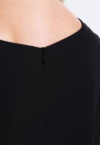 Svart klänning med detalj på baksidan som har en rundad halsringning och en dold dragkedja i nacken, med slät tyg och en minimalistisk design.