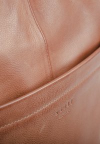 Borsa in pelle tan con superficie texture, cuciture in vista e una tasca frontale con un logo inciso discreto.