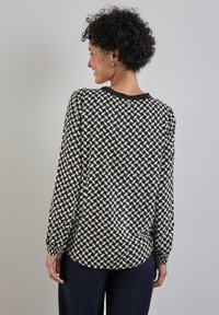 Blusa a maniche lunghe nera con un motivo geometrico beige. Tessuto morbido, scollatura rotonda e polsini arricciati. Vestibilità casual e orlo arrotondato.