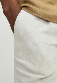 Pantalons blancs texturés avec un motif rayé subtil, partiellement recouverts par un haut beige, mettant en évidence le design de poche courbe.