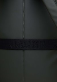 Borsa verde scuro con una superficie liscia, dotata di una tracolla nera intrecciata con la scritta "JACKIT". Presenta dettagli di cucitura visibili.