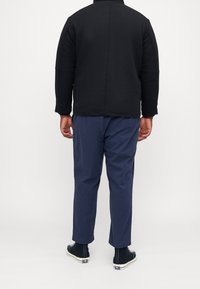 Czarna polarowa kurtka z wysokim kołnierzem, w zestawieniu z granatowymi chinosami. Buty są ciemne z białymi podeszwami. Tylny widok ukazuje detale przeszyć.
