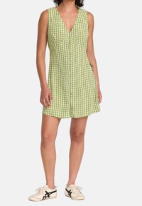 Robe à carreaux verts avec un col en V, sans manches et une fermeture à boutons sur le devant. Associée à des chaussures blanches avec des accents noirs.