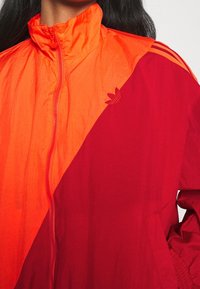 Närbild av en person som bär en ljusorange och röd Adidas zip-jacka med hög krage och Adidas trefoil-logotyp på bröstet.