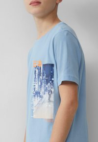 Lichtblauw katoenen T-shirt met ronde hals, voorzien van een grafische print van een stadsscene op de voorkant, geaccentueerd met oranje tekst en details.