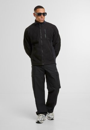 Mann trägt schwarze Fleecejacke, schwarze Cargohose, weiße Laufschuhe und schwarze Sonnenbrille, steht mit den Händen hinter dem Rücken.