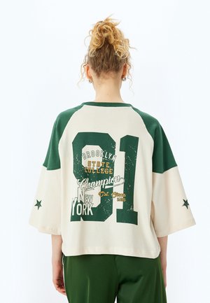 Camiseta oversized verde y crema con mangas cortas, que presenta una impresión gráfica de texto y números en la parte posterior. Tejido suave de mezcla de algodón.
