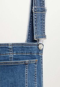 Bretelle de salopette en denim fixée au plastron avec une boucle métallique argentée et un bouton, montrant une double couture sur un tissu bleu et une poche avant.