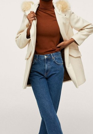 Manteau couleur crème avec un col en fourrure, associé à un pull-over marron et un jean évasé bleu. Poches et détails de couture remarquables.